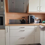 Moderne keuken met witte kasten met luxe Leonardo RVS Deluxe handgrepen van yessential.nl; een zwarte elektrische waterkoker op het fornuis, koffiezetapparaat, broodrooster en kleine apparaten op het aanrecht; een deel van een pluizige grijze kat verschijnt rechts.