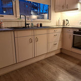 Moderne keuken met beige kasten met Leonardo RVS Deluxe handgrepen van yessential.nl, donker aanrechtblad, ingebouwde oven, waterkoker, afwasmiddel en handdoek bij de gootsteen. Grote ramen tonen 's avonds stadsgebouwen verlicht door warme lampen.