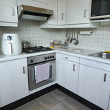Een moderne witte keuken met een Vasco RVS oven, fornuis en afzuigkap van yessential.nl met geborsteld roestvrij staal. Een friteuse en een pan staan op het aanrecht, keukengerei hangt erboven en een gestreepte handdoek hangt aan vierkante handgrepen.