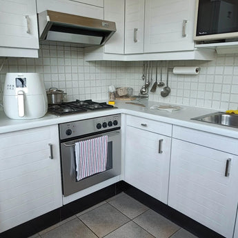 Een moderne witte keuken met een Vasco RVS oven, fornuis en afzuigkap van yessential.nl met geborsteld roestvrij staal. Een friteuse en een pan staan op het aanrecht, keukengerei hangt erboven en een gestreepte handdoek hangt aan vierkante handgrepen.