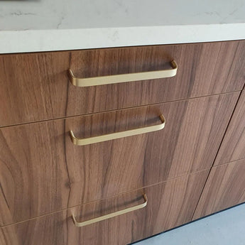 Een close-up van drie houten lades met de luxe handgreep keuken Leonardo Gold Deluxe van yessential.nl in goud, afgewerkt met een wit aanrechtblad. Het hout vertoont een natuurlijk nerfpatroon.