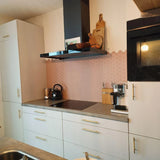Een moderne keuken met witte kastjes, Remo Gold Deluxe handgrepen van yessential.nl, een zwart fornuis en waterkoker, plus een koffiezetapparaat. De roze zeshoekige tegelachterwand voegt charme toe, terwijl luxe keuken grepen de stijlvolle look afmaken.