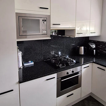 Moderne keuken met witte kasten, moderne handgrepen en de Arriero Black Deluxe glanzend zwarte backsplash van yessential.nl voor een strakke touch. Kenmerken zijn onder andere een zwart granieten aanrechtblad, ingebouwde magnetron, roestvrijstalen oven, gasfornuis en waterkoker.