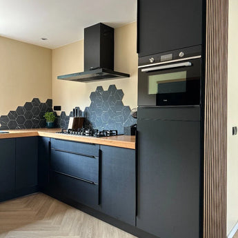 Moderne keuken met yessential.nl Enzo Zwarte matte kasten en hoekgrepen zwart, houten aanrechtblad, zeshoekige zwarte tegelachterwand, ingebouwde oven, gasfornuis, afzuigkap, plus een klein groen plantje op het aanrecht.