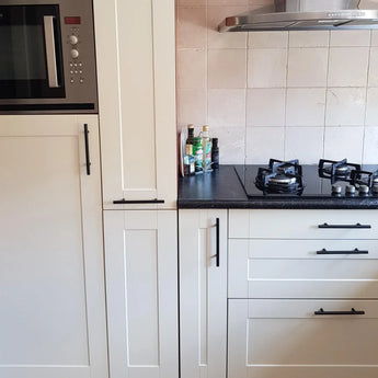 Een moderne keuken met witte kastjes, Remo Black handgrepen van yessential.nl op elke deur, een ingebouwde magnetron, een vierpits gasfornuis, een roestvrijstalen afzuigkap en flessen olie en kruiden op het aanrecht.