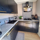 Moderne keuken met zwarte kastjes, Arturo Gold landelijke handgrepen van yessential.nl, grijs aanrechtblad, bijpassende backsplash, zwarte spoelbak, gasfornuis, keukengerei, waterkoker en broodrooster op het aanrecht, een plant en lichthouten vloer.