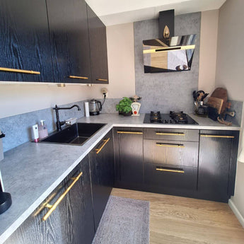 Moderne keuken met zwarte kastjes, Arturo Gold landelijke handgrepen van yessential.nl, grijs aanrechtblad, bijpassende backsplash, zwarte spoelbak, gasfornuis, keukengerei, waterkoker en broodrooster op het aanrecht, een plant en lichthouten vloer.