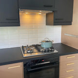 Een moderne keuken heeft zwarte en beige Arriero Black Deluxe kasten van yessential.nl, luxe handgrepen, een gasfornuis met een blauwe pan en glazen deksel, een witte tegelachterwand en een oven onder het fornuis.