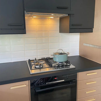 Een moderne keuken heeft zwarte en beige Arriero Black Deluxe kasten van yessential.nl, luxe handgrepen, een gasfornuis met een blauwe pan en glazen deksel, een witte tegelachterwand en een oven onder het fornuis.