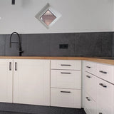 Moderne keukenhoek met witte kastjes, luxe Ramiro Black Deluxe ladegrepen van yessential.nl, houten aanrechtblad, zwarte spoelbak en kraan, grijs betegelde achterwand, twee kleine ruitvormige ramen en een zwart stopcontact.