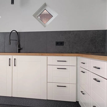 Moderne keukenhoek met witte kastjes, luxe Ramiro Black Deluxe ladegrepen van yessential.nl, houten aanrechtblad, zwarte spoelbak en kraan, grijs betegelde achterwand, twee kleine ruitvormige ramen en een zwart stopcontact.