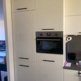 Moderne keuken met witte kasten en Marano Black handgrepen van yessential.nl, een ingebouwde oven die 10:15 weergeeft, plus een aanrechtblad, gootsteen en woonkamer met tv zichtbaar op de achtergrond.