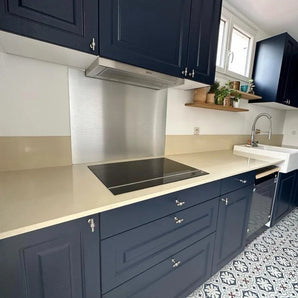 Moderne keuken met marineblauwe kastjes met Rando RVS knop van yessential.nl, beige aanrechtblad, elektrische kookplaat, roestvrijstalen achterwand, boerengootsteen, tegelvloer met patroon en planken boven met potplanten en keukengerei.