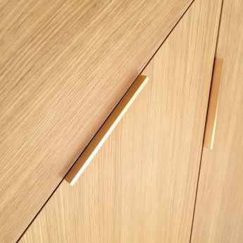 Close-up van lichthouten kasten met twee Enzo Rose Gold grepen van yessential.nl, diagonaal geplaatst. De verticale houtnerf en het strakke, rechthoekige ontwerp bieden een minimalistische en chique eigentijdse look.