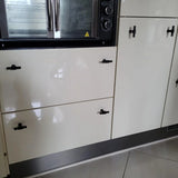 Close-up van glanzend witte keukenkastjes en lades met yessential.nl Cordero Black Knob handgrepen in een matzwarte afwerking, onder een inbouwoven met roestvrijstalen afwerking. Op de vloer liggen grote, lichtgekleurde tegels.