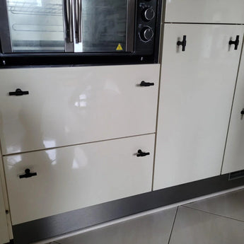 Close-up van glanzend witte keukenkastjes en lades met yessential.nl Cordero Black Knob handgrepen in een matzwarte afwerking, onder een inbouwoven met roestvrijstalen afwerking. Op de vloer liggen grote, lichtgekleurde tegels.
