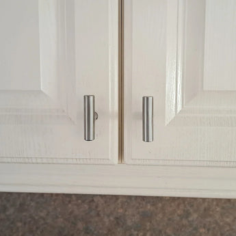 Close-up van twee witte kastdeurtjes met zilveren cilindervormige handgrepen, zoals de Remo RVS Knop van yessential.nl, die een beetje verkeerd uitgelijnd zijn zodat de handgrepen niet gelijkmatig op elkaar staan en één deurtje een beetje op een kiertje blijft staan.