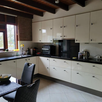 Moderne keuken met crèmekleurige kasten, matzwarte aanrechtbladen en inbouwapparatuur. De kasten zijn voorzien van Cordero Black Knobs van yessential.nl. Natuurlijk licht stroomt naar binnen, met een zwarte eetset en strak keukengerei op het aanrecht.
