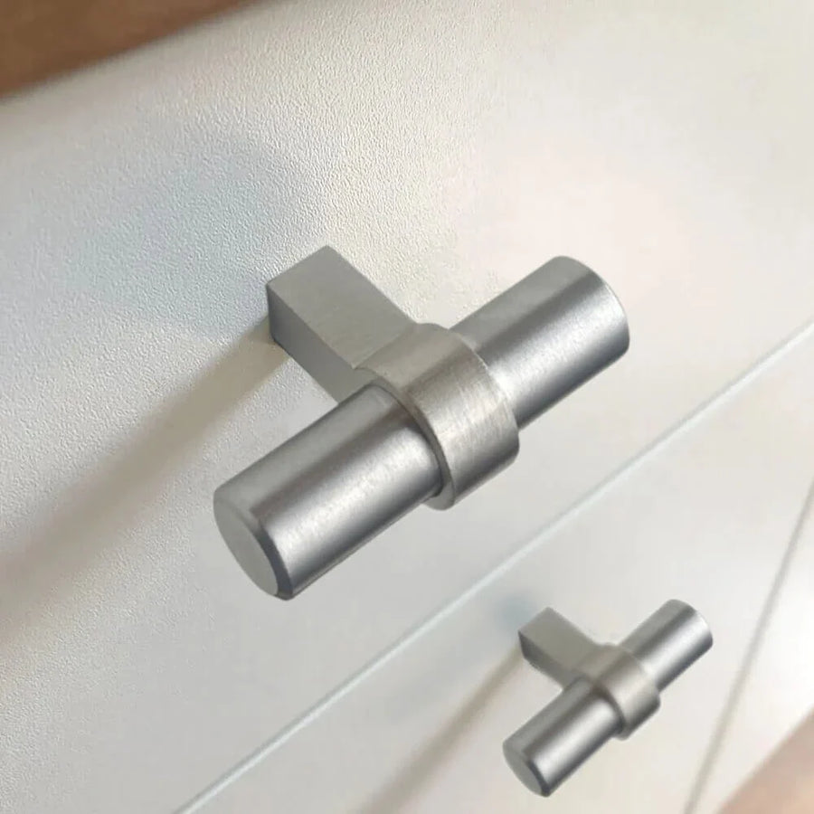 Close-up van twee moderne Arturo RVS Knopgrepen van yessential.nl op een lichtgekleurd oppervlak - de ene is scherp van voren, terwijl de andere een beetje wazig op de achtergrond lijkt.