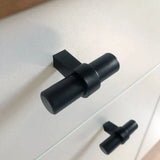 Close-up van twee Arturo zwarte knoppen van yessential.nl in een moderne T-knop keukenstijl, bevestigd op een witte kast. Het cilindrische ontwerp heeft een horizontale staaf op een verticale basis voor een strakke, eigentijdse look.