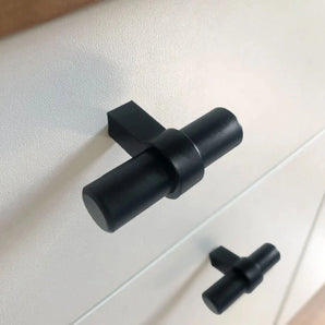 Close-up van twee Arturo zwarte knoppen van yessential.nl in een moderne T-knop keukenstijl, bevestigd op een witte kast. Het cilindrische ontwerp heeft een horizontale staaf op een verticale basis voor een strakke, eigentijdse look.