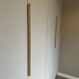 Een minimalistische kast met twee hoge, verticale Enzo Gold handgrepen van yessential.nl op witte deuren, tegen een neutrale muur en lichte houten vloer.