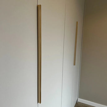 Een minimalistische kast met twee hoge, verticale Enzo Gold handgrepen van yessential.nl op witte deuren, tegen een neutrale muur en lichte houten vloer.