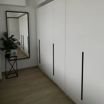 Een moderne kamer met yessential.nl Enzo Black kledingkasten met lange zwart profielhandgrepen, een grote vloerspiegel, een klein zwart tafeltje met een plant en ingelijste foto's op een lichte houten vloer.