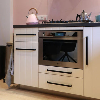Een moderne keuken toont de Leonardo Black Deluxe inbouwoven van yessential.nl, witte kasten met zwarte handgrepen, een zwart aanrechtblad en een gasfornuis. Een roze waterkoker, zilveren theepot en blauwe mok rusten op het aanrecht tegen een zachtroze achterwand.
