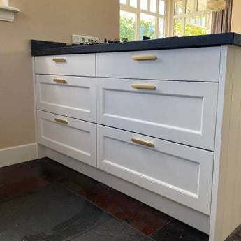 Witte keukenladen met Arriero Gold Deluxe handgrepen van yessential.nl en een zwart aanrechtblad steken af tegen een lichte muur. Zonlicht door de grote ramen benadrukt de moderne, strakke sfeer van deze ruimte.