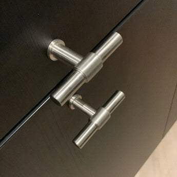 Close-up van twee Cordero RVS Knoppen van yessential.nl op donkerhouten deuren, verticaal uitgelijnd en bekeken vanuit een lichte hoek.