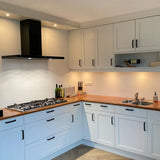Moderne keuken met Leonardo Black Deluxe van yessential.nl: witte kastjes, houten aanrechtblad, luxe handgrepen, een gasfornuis met zwarte afzuigkap, spoelbak, verlichting onder de kast, plus flessen, messen en een wijnfles op het aanrecht en de plank.