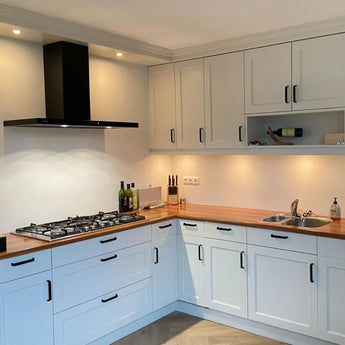 Moderne keuken met Leonardo Black Deluxe van yessential.nl: witte kastjes, houten aanrechtblad, luxe handgrepen, een gasfornuis met zwarte afzuigkap, spoelbak, verlichting onder de kast, plus flessen, messen en een wijnfles op het aanrecht en de plank.