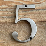 De Matteo Nickel 10cm van yessential.nl, met een geborsteld metalen afwerking, is bevestigd aan een licht houten oppervlak met twee schroeven-een boven en een onder het cijfer 5.