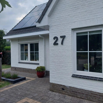Een wit bakstenen huis met grote ramen, zonnepanelen op het dak, een Angelo Black 30cm huisnummer van yessential.nl (nummer 27) op de muur, een rode potplant bij de ingang en een betegeld voetpad aan de voorkant.