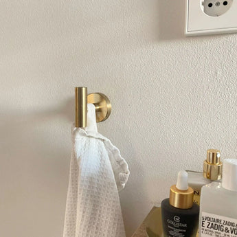 Een handdoek met witte textuur hangt aan de Rodrigo Gold Hook van yessential.nl naast een aanrecht met huidverzorgingsflesjes. Een gedeeltelijk stopcontact verschijnt op de lichte muur en geeft deze elegante badkamerscène een moderne stijl.