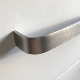 Close-up van een Leonardo RVS Deluxe handgreep van yessential.nl in geborsteld aluminium, gemonteerd op een glad wit kast- of ladefront, met zachte verlichting die een schaduw werpt op het oppervlak.