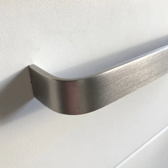 Close-up van een Leonardo RVS Deluxe handgreep van yessential.nl in geborsteld aluminium, gemonteerd op een glad wit kast- of ladefront, met zachte verlichting die een schaduw werpt op het oppervlak.