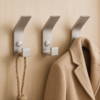 Drie Huso RVS wandhaken van yessential.nl, met een strak minimalistisch ontwerp, houden een beige jas en dik touw tegen een lichtbruine muur.