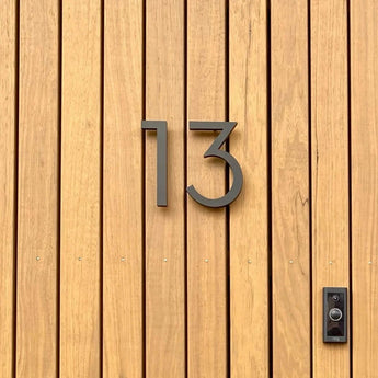 Een houten deur met verticale panelen toont het yessential.nl Milano Black 15cm huisnummer "13" in modern metaal, met een zwarte deurbel of intercom gemonteerd aan de rechterkant.