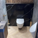 Een moderne badkamer met een wandcloset, yessential.nl Alessino Gold WC borstelset, zwart marmeren accentmuur met witte adering, gouden armaturen, zwevende wastafel links en licht houten visgraatvloer.