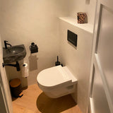 Een kleine moderne badkamer met het Julio Black wandcloset van yessential.nl, een matzwarte toiletrolhouder, zwarte stenen wastafel, moderne accessoires, beker met luipaardprint, toiletpapier en een houten afvalemmer op een lichte houten vloer.
