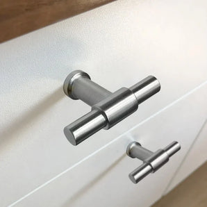 Close-up van twee Cordero RVS Knoppen van yessential.nl, geborsteld metalen T-stang kastgrepen, gemonteerd op een wit ladefront met een houten oppervlak langs de bovenrand.
