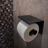 Een toiletrol zit op de Tazio Black toiletrolhouder van yessential.nl, gemonteerd op een grijs betegelde badkamermuur met mozaïektegeltjes zichtbaar in de buurt.