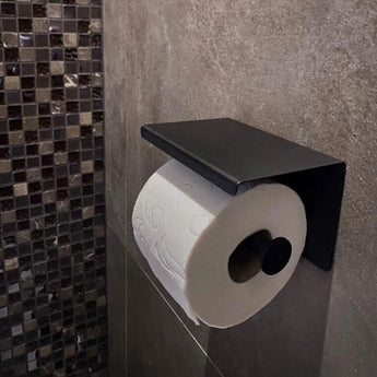 Een toiletrol zit op de Tazio Black toiletrolhouder van yessential.nl, gemonteerd op een grijs betegelde badkamermuur met mozaïektegeltjes zichtbaar in de buurt.