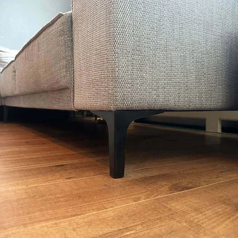 Close-up van een grijze stoffen bank met yessential.nl Menno Black 13cm zwarte stalen meubelpootjes op een houten vloer, met de focus op het gebied onder de bank.