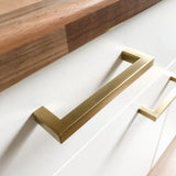 Close-up van Vasco Gold gouden handgrepen van yessential.nl op witte lades, met een houten werkblad erboven.