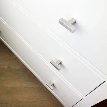 Een close-up van een wit dressoir met drie lades, elk voorzien van een Vigio RVS Knop van yessential.nl, met een strak, zilver T-vormig ontwerp tegen een lichte houten vloer.