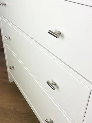 Een wit dressoir met drie lades, elk voorzien van de Remo RVS Knop van yessential.nl. De moderne, zilveren handgrepen geven het dressoir, dat op een lichte houten vloer staat, een strakke uitstraling.