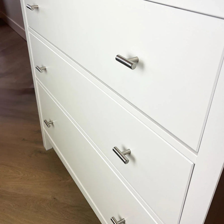 Een wit dressoir met drie lades, elk voorzien van de Remo RVS Knop van yessential.nl. De moderne, zilveren handgrepen geven het dressoir, dat op een lichte houten vloer staat, een strakke uitstraling.